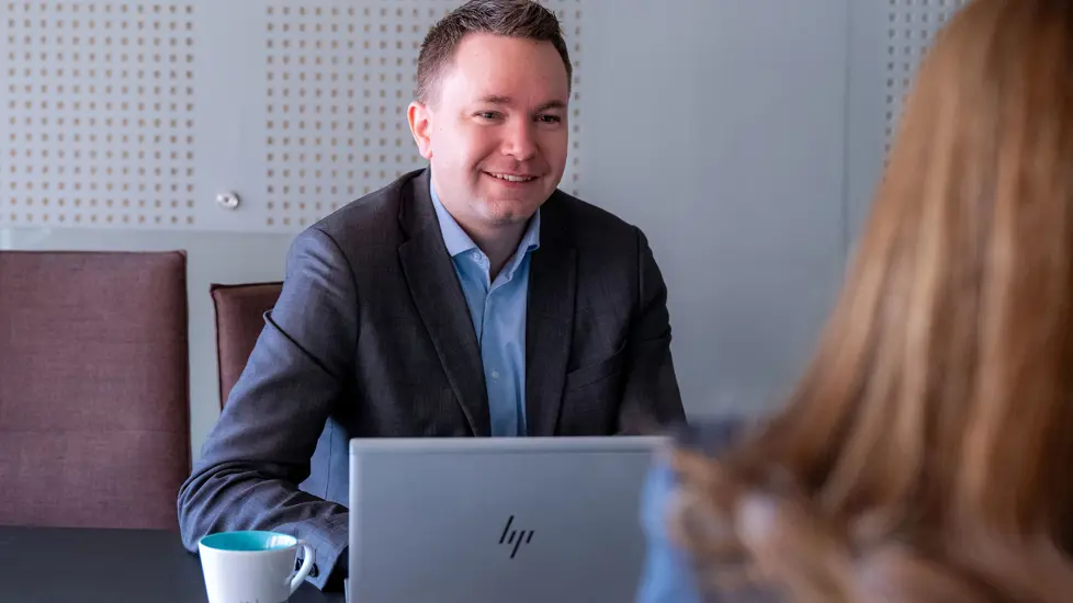 Mann i skjorte og dress med PC foran seg på bordet, i samtale med person med bakhodet mot kamera.