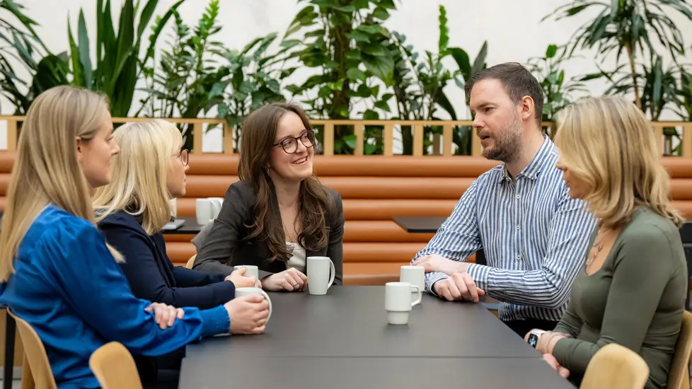 Fem kollegaer, fire kvinner og en mann, sitter og snakker sammen rundt et bord.